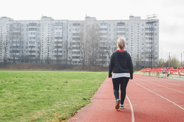 Middle-aged woman practicing race walking in a street stadium, back view. Active lifestyle, weight loss concept