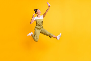 Photo of funky lady jump hold phone make selfie show v-sign wear green overall sneakers isolated yellow background