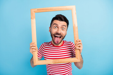 Photo of funny fooling young guy wear red outfit holding looking wooden frame showing tongue isolated blue color background