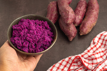 Hand holding of mashed purple sweet potatoes in a bowl with purple yams pile and a cloth placed on a dark brown background. Space for text. Vintage background. Healthy fruits concept