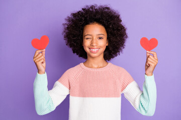 Photo portrait of small schoolgirl showing two little red paper hearts winking isolated purple color background