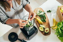 Woman is preparing a healthy detox drink in a blender - a  green smoothie with fresh fruits, green spinach and avocado. Healthy eating concept, ingredients for smoothies on the table, top view