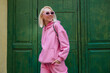 Happy smiling fashionable woman wearing trendy pink sport chic style outfit, sunglasses, posing in street on green background. Glasses, pink hoodie, trousers. Spring fashion concept. Copy, empty space