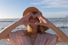 Caucasian Woman Wearing Hat Making Heart Shape Looking At Camera And Smiling At The Beach