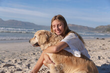 Caucasian Woman Sitting On Sand Embracing A Dog At The Beach
