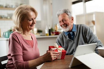 Wall Mural - Senior couple having video call. Happy husband giving his wife a gift..