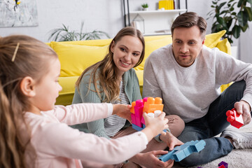 Sticker - Positive parents looking at kid playing building blocks on blurred foreground.