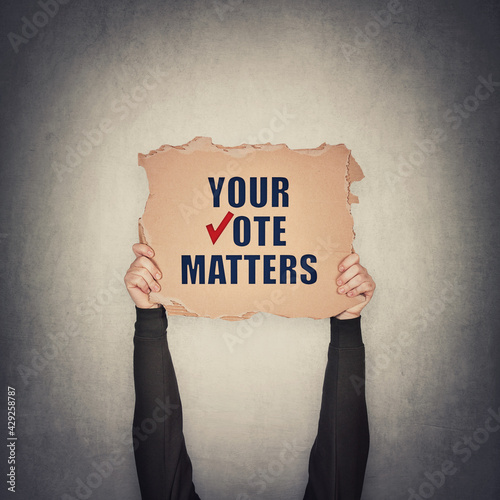 Activist hands holding up a cardboard banner with text message your vote matters, isolated on gray wall background. People legal and democratic rights, every voice counts. Election campaign agitation