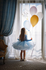 Little Caucasian girl in babydoll dress standing by big french window in bedroom holding baloons in hands and looking outside at the sea. Birthday concept. Image with selective focus