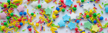 Banner With Multicolor Festive Background Of A Scattering Of Sugar Candy Sprinkles For Cupcakes And Other Pastries In Form Of Stars, Sticks And Balls. Festive Background Of Red, Blue, Yellow And Green
