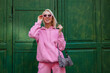 canvas print picture - Street style, fashion portrait of happy smiling woman wearing trendy pink sport chic style outfit with hoodie, sunglasses, zebra print bag, posing in street on green background. Copy, empty space
