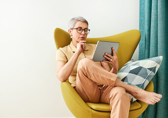 Wall Mural - Thoughtful mature woman with closed eyes sitting at home holding a digital tablet