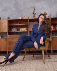 Wall Mural - Young beautiful pretty woman in blue suit sitting in green armchair in bright interior with crossed legs and looking at you or in camera