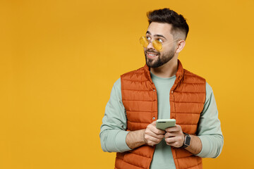 Wall Mural - Young smiling happy friendly caucasian man 20s wear orange vest mint sweatshirt glasses using mobile cell phone look aside isolated on yellow background studio portrait. Technology lifestyle concept.