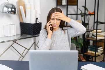 Worried young business woman holding head in despair, looks at the laptop screen and talks on the phone, tired and feels stress