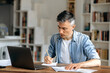 © Kateryna - Focused smart gray-haired manager, coach or freelancer dressed in casual stylish clothes, working at home with laptop and documents at the table, preparing a business project, taking notes