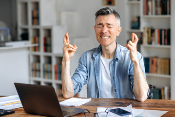 Mature gray-haired caucasian, male freelancer, top manager, in stylish clothes, works at a laptop, sits at work desk, worries, hoping for the desired result, gestures with his hands, closes his eyes