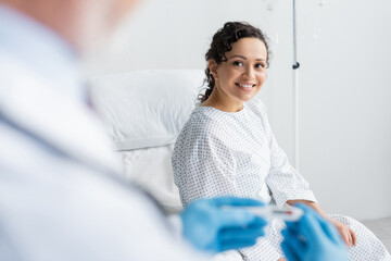 african american woman smiling near doctor in latex gloves holding thermometer on blurred foreground