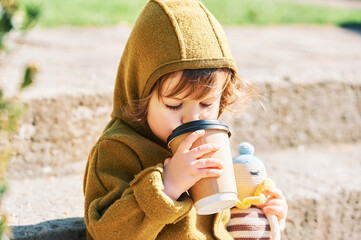 Funny little kid drinking from takeaway coffe cup in early morning, sitting on stairs in public park