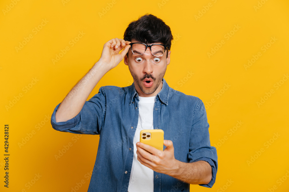 Amazed shocked caucasian guy holding smartphone in his hand, looking at the phone in surprise with his glasses raised, stunned facial expression, stands on isolated orange background - obrazy, fototapety, plakaty 
