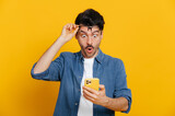 Fototapeta  - Amazed shocked caucasian guy holding smartphone in his hand, looking at the phone in surprise with his glasses raised, stunned facial expression, stands on isolated orange background