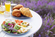 Close up view of wooden table decorated in lavender field. Delicious croissants, glasses of orange juice, honey jar and fresh plates with eggs, salmon and salad and vase with lavender bouquet.