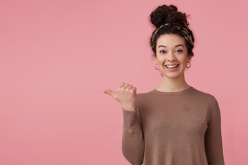 Happy woman with dark curly hair bun. Wearing headband, earrings, brown sweater. Has make up. Watching at the camera and pointing thumb to the left at copy space, isolated over pastel pink background