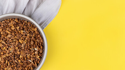 Wall Mural - Organic homemade roasted granola cereal with oats, flax, almonds, chocolate in round baking tray with linen textile on bright yellow background. Top view, flat lay muesli wide banner with copy space