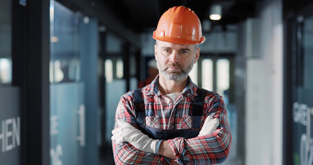 Wall Mural - Caucasian engineer bearded adult crossing arms for camera posing in protective bodysuit operating in modern building interior. Electrician. Jobs concept.