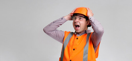 Wall Mural - young engineer architect clutching his head in helmet on grey studio background, foreman shocked