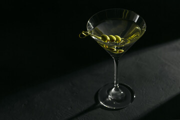 Glass of classic dry martini cocktail with olives on dark stone table against a black background.