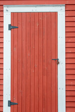 A Large Vintage Red Wooden Shed Or Barn Door With White Trim On An Outcrop Building.  The Wood Door Is Closed And Has Two Hinges On The Left Side With An Old Latch On The Right Side. 