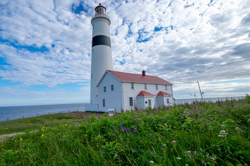 Point Amour Historic Lighthouse on the edge of the Atlantic Ocean. The blue sky is hidden by fluffy clouds.A building with two entrances and a red roof is joined to tower which has a large lamplight.