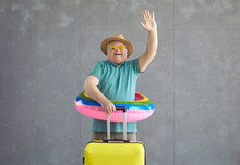 Happy Mature Tourist Leaving For Vacation Alone. Excited Fat Senior Man In Sun Hat, Pool Lifesaver And Cool Sunglasses Holding Suitcase And Waving Goodbye As He's Going On Travel Adventure Far Away
