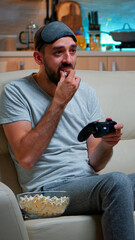 Wall Mural - Upset pro gamer sitting on couch and playing soccer videogames for online competition using gaming joystick. Disappointed man looking at television eating popcorn late at night in kitchen