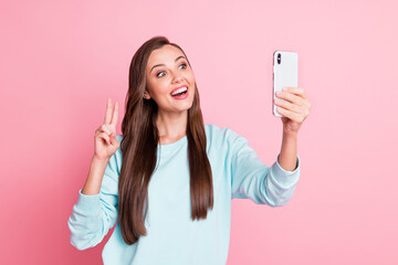 Sticker - Photo of attractive charming young lady wear blue sweater hold phone show v-sign selfie isolated on pink color background