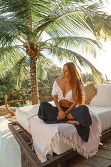 Wall Mural - Woman is relaxing on a luxurious sunbed under a palm tree in lotus position on a tropical beach near swiming pool