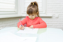 Little Caucasian Girl In Red Clothes Sits At A Table In A White Room And Draws