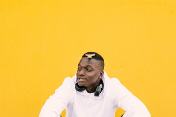 Wall Mural - Young african American black man combing his afro hair while seated on a yellow wall background, wearing a white sweatshirt and wireless headphones. Diversity concept. Copy space