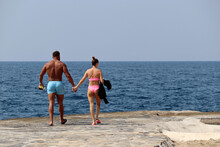 Couple Walking On A Stone Beach On Sea Background. Muscular Tanned Man In Blue Trunks And Girl With White Skin In Pink Bikini Together, Romantic Holiday And Vacation