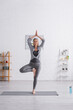 © LIGHTFIELD STUDIOS - middle aged woman in sportwear standing in tree pose on yoga mat at home