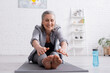 © LIGHTFIELD STUDIOS - happy mature woman with grey hair stretching on yoga mat near sports bottle
