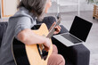 © LIGHTFIELD STUDIOS - blurred middle aged woman learning to play acoustic guitar near laptop with blank screen on sofa