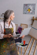 © LIGHTFIELD STUDIOS - mature woman holding paintbrush and laptop while looking at canvas