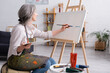 © LIGHTFIELD STUDIOS - cheerful middle aged woman holding paintbrush and palette while painting on canvas
