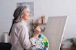 © LIGHTFIELD STUDIOS - pleased middle aged woman holding paintbrush and palette with colorful paints near blank canvas
