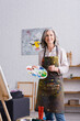 © LIGHTFIELD STUDIOS - positive middle aged woman holding paintbrush and palette with colorful paints near easel