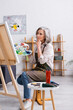 © LIGHTFIELD STUDIOS - pensive mature artist holding paintbrush and palette while sitting near canvas