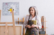© LIGHTFIELD STUDIOS - pleased middle aged artist in apron with spills holding paintbrush and looking at canvas on easel
