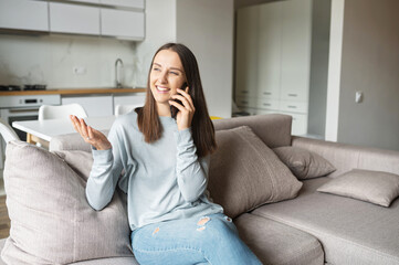 Wall Mural - Charming young woman talks on the smartphone sitting on the couch at home, gossips with friends. Serene housewife in casual wear sits on the comfortable couch and holding phone conversation